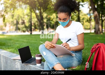 Black Student Girl prenant des notes portant un masque de visage apprendre à l'extérieur Banque D'Images