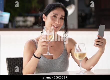 Femme adulte assise dans un restaurant en plein air prenant un selfie avec son téléphone tout en tenant un verre de vin regardant l'appareil photo Banque D'Images