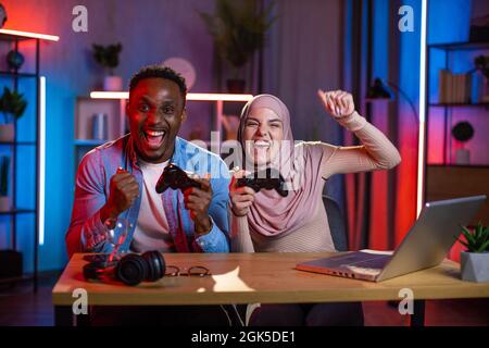 Joyeux homme africain et femme musulmane se sentant heureux tout en gagnant dans les jeux vidéo sur ordinateur portable. Couple multi-ethnique appréciant le divertissement à la maison pendant la soirée. Banque D'Images