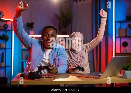 Homme noir joyeux et femme arabe se sentant heureux tout en gagnant dans les jeux vidéo sur ordinateur portable. Couple multi-ethnique appréciant le divertissement à la maison pendant la soirée. Banque D'Images