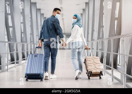 Joyeux couple islamique en masques médicaux à pied avec des valises à l'aéroport Banque D'Images