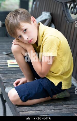 Un petit garçon triste est assis sur un banc de parc. Banque D'Images