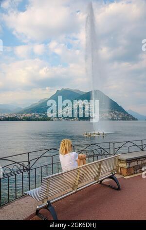 Femme regardant le jet d'eau de la fontaine Paradiso de la ville de Lugano dans le lac de Lugano en Suisse. Banc au bord de l'eau du lac de Lugano avec Monte San Banque D'Images