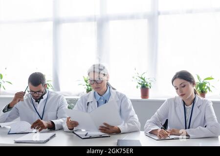 médecin asiatique d'âge moyen regardant dans un dossier papier près de jeunes collègues pendant la réunion Banque D'Images