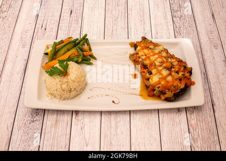 Aubergine farcie de légumes et gratin de fromage avec sauce à la carotte et garniture de haricots verts cuits avec du riz blanc sur un plateau blanc Banque D'Images