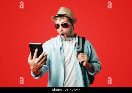 Un homme dans un chapeau se tient sur un fond rouge et tient une tablette dans sa main. Il regarde avec émotion de surprise. Studio portrait d'un voyageur dans des vêtements décontractés. Banque D'Images