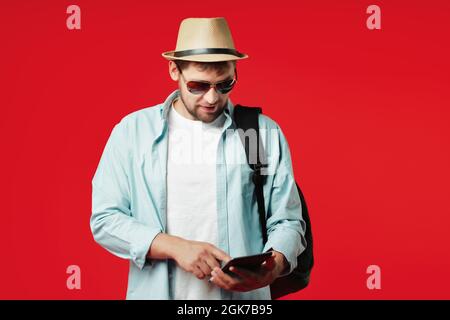 Un touriste caucasien dans un chapeau et des lunettes de soleil avec un sac à dos sur son épaule se tient sur un fond rouge et regarde l'application sur la tablette. Le concept d'une application mobile pour les voyageurs. Banque D'Images