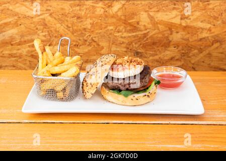 Délicieux hamburger de bœuf avec une tranche de fromage de chèvre, confiture de tomates, oignon croustillant, feuilles d'épinards, pain aux graines et une portion de frites Banque D'Images