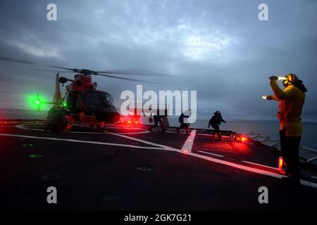 Env. Ryan Dunkle, un membre de l’équipage de la Garde côtière Healy (WAWGB 20), donne des ordres à une équipe d’arrimage lors d’opérations de formation en vol conjointes de nuit avec la station aérienne Kodiak au large de la côte de l’Alaska pendant le déploiement de Healy dans l’Arctique le 25 août 2021. Les agents de signalisation d'atterrissage sont responsables de communiquer avec l'équipage et les membres d'équipage de la découpeuse pendant les opérations de vol. Banque D'Images