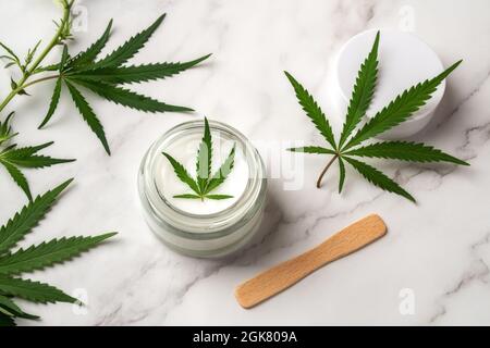 Crème hydratante de soin de la peau avec feuille de cannabis sativa verte dans un pot en verre et tige de chanvre frais et feuilles sur la surface de marbre. Cosmétiques pour la marijuana. Banque D'Images