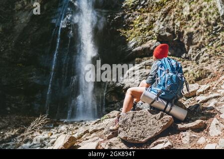 Femme avec un sac à dos en chapeau rouge vêtu de vêtements de randonnée actifs assis près de la cascade de la rivière de montagne et appréciant la puissance de nature éclabousse. Banque D'Images