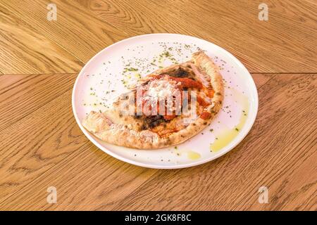 Recette typique de calzone italienne avec sauce tomate, fromage râpé, origan séché et huile d'olive sur plaque blanche et table en bois Banque D'Images