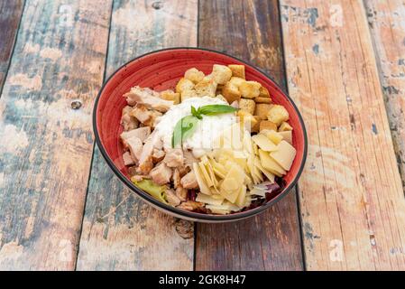 Salade césar incroyable avec beaucoup de parmesan grillé, morceaux de poulet, mayonnaise et croûtons à l'intérieur d'un bol rouge sur fond de bois Banque D'Images