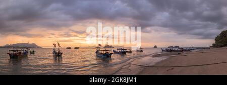 Large angle de bateaux de pêche avec drapeaux nationaux sur la côte de sable humide lavée par la mer sous un ciel sombre et nuageux au coucher du soleil en Malaisie Banque D'Images