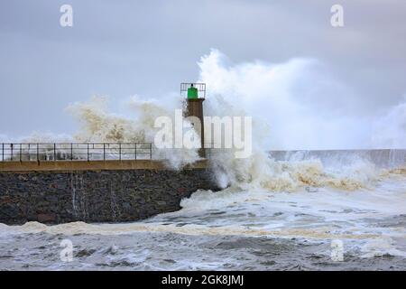 D'énormes vagues de mer mousseuse s'écrasant contre le brise-lames de pierre avec l'ancienne tour de phare contre le ciel bleu ciel nuageux dans le port de Viavelez dans les Asturies Espagne Banque D'Images