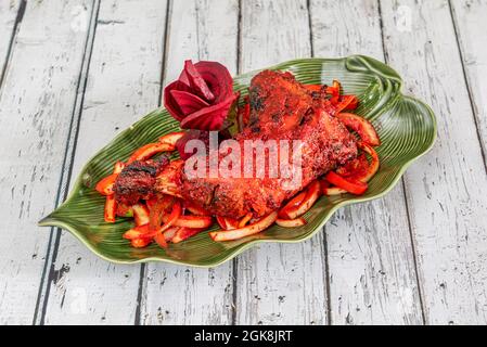 Cuisse de poulet Tandoori assaisonnée avec beaucoup de sauce rouge typique indienne sur une belle assiette en forme de feuille verte sur une table blanche Banque D'Images