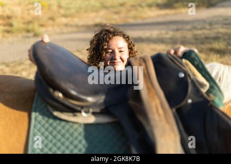 Femelle d'âge moyen avec des cheveux bouclés mettant selle sur l'arrière de jument sur la ferme en plein soleil Banque D'Images