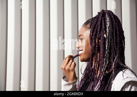 Vue latérale d'une jeune femme afro-américaine élégante et heureuse avec des tresses afro-violettes teintées appliquant un rouge à lèvres violet tout en se tenant contre le wal à rayures grises Banque D'Images