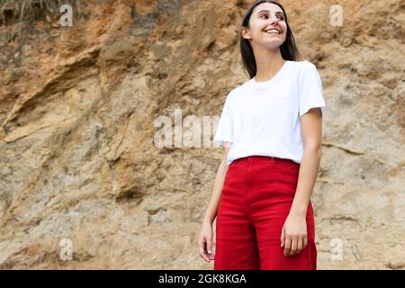 Jeune femme jeune et heureuse contemplative, en t-shirt blanc et en jeans rouges, regardant loin tout en se tenant debout sur une terre difficile contre le support Banque D'Images