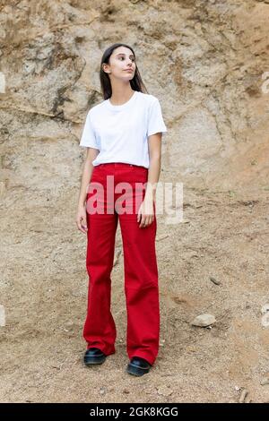 Jeune adolescente contemplative en t-shirt blanc et jeans rouges regardant loin tout en se tenant debout sur un terrain accidenté contre le support Banque D'Images