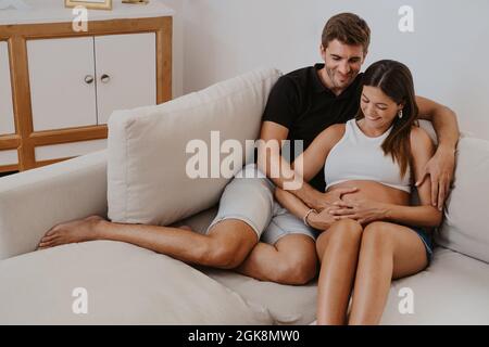 homme embrassant le ventre d'une femme enceinte bien-aimée tout en se reposant sur un canapé dans le salon Banque D'Images