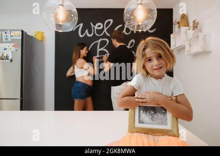 Charmant enfant avec futur frère ultrasons image regardant l'appareil photo contre la mère enceinte et le père écrire sur tableau noir dans la maison Banque D'Images