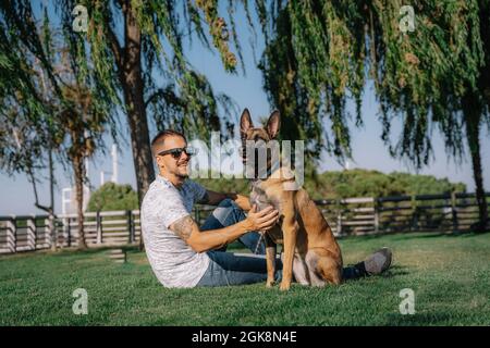 Vue latérale d'un homme tatoué dans des lunettes de soleil avec des Malinois reposant sur la prairie et regardant l'un l'autre Banque D'Images