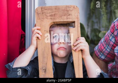 Enfant étonné dans des lunettes de sécurité avec pièce en bois regardant l'appareil photo contre la récolte non reconnaissable papa en lumière du jour Banque D'Images