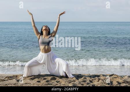 Jeune femme ethnique aux bras levés qui se squat sur la côte sablonneuse tout en pratiquant le yoga contre la mer en plein soleil Banque D'Images