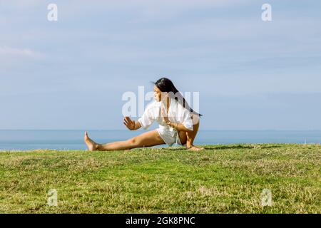 Les jeunes femmes ethniques qui se disputent à Skandasana posent tout en pratiquant le yoga sur la côte de l'herbe contre la mer sous un ciel nuageux Banque D'Images