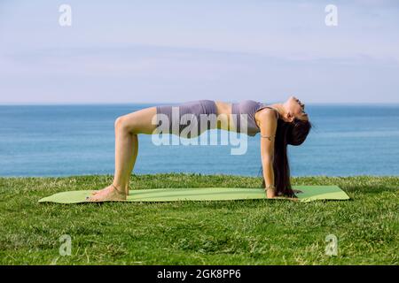 Vue latérale de la jeune femme ethnique debout dans Ardha Purvottanasana pose tout en pratiquant le yoga sur tapis contre l'océan Banque D'Images