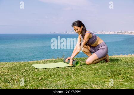 Contenu jeune femme asiatique dans des vêtements de sport roulant tapis de yoga sur la côte d'herbe contre l'océan sans fin dans la lumière du soleil Banque D'Images