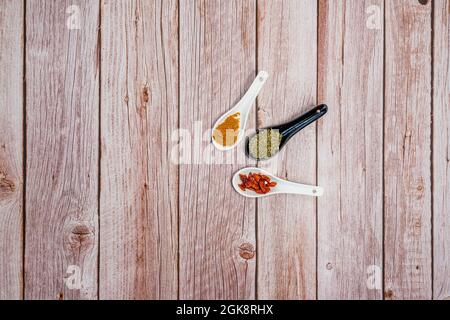 trois cuillères en porcelaine, une noire et deux blanches avec cayenne entière, curry jaune et feuilles d'origan séchées sur une table en bois Banque D'Images