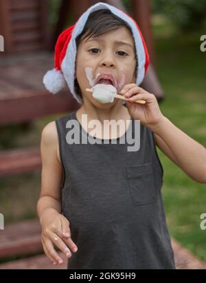 Brunette petit garçon savoure une crème glacée à l'heure de Noël portant un chapeau rouge du Père Noël et regardant l'appareil photo. Vertical. Argentine Banque D'Images