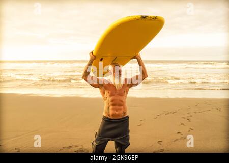 Happy FIT senior ayant du plaisir à surfer à l'heure du coucher du soleil. Entraînement sportif d'homme barbu avec planche de surf sur la plage. Personnes âgées en bonne santé mode de vie et extr Banque D'Images