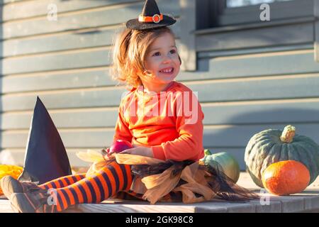 Une jolie petite fille 2-3 dans un costume de sorcière orange et noir d'Halloween, se trouve à côté de citrouilles sur la terrasse d'une maison en bois gris Banque D'Images