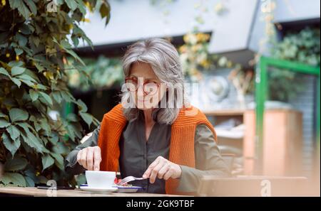 Une femme âgée aux cheveux gris mange un délicieux dessert assis à table à l'extérieur Banque D'Images