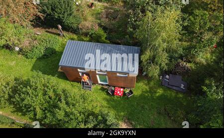 07 septembre 2021, Hessen, Schöneck-Kilianstädten: Uwe Christof se tient à l'entrée de sa petite maison, qui est située dans son jardin d'allotement (vue aérienne prise avec un drone). Depuis des années, le spécialiste INFORMATIQUE de la retraite anticipée fait campagne pour ce qu'on appelle les petites maisons, essentiellement transportables qui, malgré leur faible encombrement, sont censées offrir à leurs habitants tout ce dont ils ont besoin pour vivre et peuvent être mis en place partout où cela est permis et les propriétaires aiment cela. À Hesse aussi, cela pourrait se transformer en une forme de vie pour l'avenir. (À dpa 'petites maisons avec un grand avenir? Banque D'Images