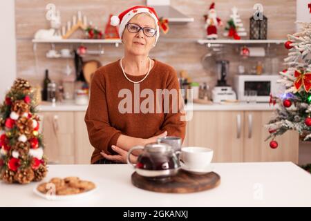 Portrait de femme âgée avec chapeau de père noël debout à table dans la cuisine décorée de Noël appréciant la saison d'hiver. Grand-mère célébrant les vacances de noël. Biscuits maison traditionnels sur la table Banque D'Images