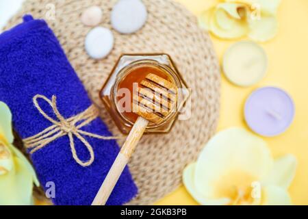 Soin spa au miel. Miel d'or dans un pot, fleurs d'orchidées, serviettes et bougies parfumées. Soin naturel de la peau à la maison. Arrière-plan jaune, vue de dessus. Banque D'Images