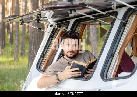 Beau jeune homme lisant le livre assis par la fenêtre ouverte de la maison sur roues Banque D'Images