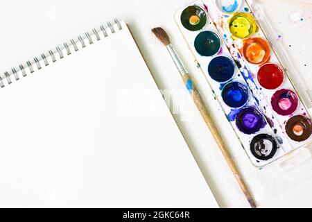 Le carnet d'esquisses ouvert, la feuille blanche de papier aquarelle, les peintures multicolores et le pinceau se trouvent sur la vue du dessus de la table. Leçon de mise en plan. Éducation art. Retour à s. Banque D'Images