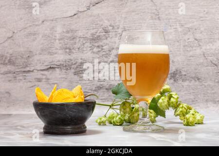 Verre de bière avec une branche de houblon et un bol avec des chips de pomme de terre sur fond gris. Banque D'Images