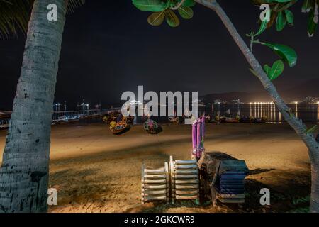 Hamacs de plage empilés sur le sable d'une plage thaïlandaise avec une petite jetée et une multitude de bateaux amarrés à longue queue Banque D'Images