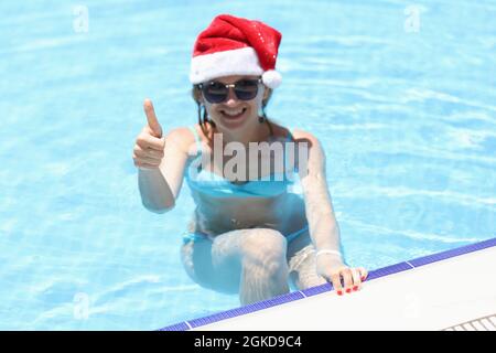 Jeune femme en lunettes de soleil et chapeau du père noël nageant dans la piscine et montrant le pouce vers le haut Banque D'Images