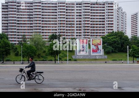 Photographie de rue à Pyongyang, un cycliste avec des affiches de propagande et un bloc de logement derrière lui.Une route large, pas de voitures. Banque D'Images