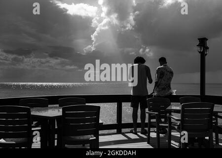 Deux amis se tenant sur une terrasse en bois, profitant de la vue sur la mer, après un épisode pluvieux. Le soleil pière les nuages. Banque D'Images