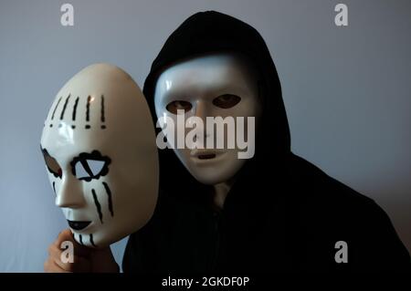 Homme à capuche noire portant un masque blanc anonyme et tenant un masque blanc féminin sur fond gris Banque D'Images