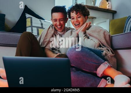 Profitez des meilleurs spectacles en ligne ensemble. Un jeune couple de reines heureux riant joyeusement tout en regardant leur émission préférée sur un ordinateur portable. Jeune LGBTQ+ co Banque D'Images