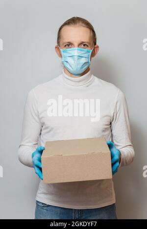 Livraison en toute sécurité, jeune messager pendant la quarantaine du coronavirus. Homme tenant une boîte en papier avec de la nourriture, ou des marchandises dans des gants médicaux et un masque de protection. Banque D'Images